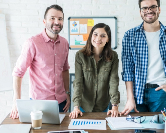 Três profissionais sorrindo em um escritório, atrás de uma mesa com laptops e gráficos.