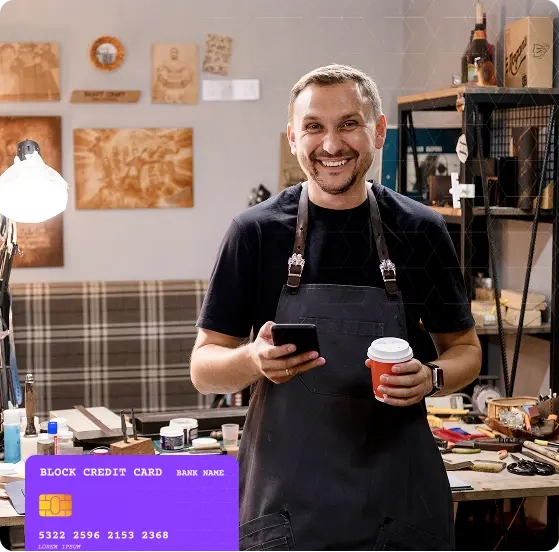 Homem sorridente em oficina artesanal segurando um celular e um café, com ferramentas ao fundo e uma ilustração de cartão de crédito sobreposta no canto.