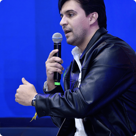 Homem palestrando em um palco, usando roupa social e expressando postura confiante.